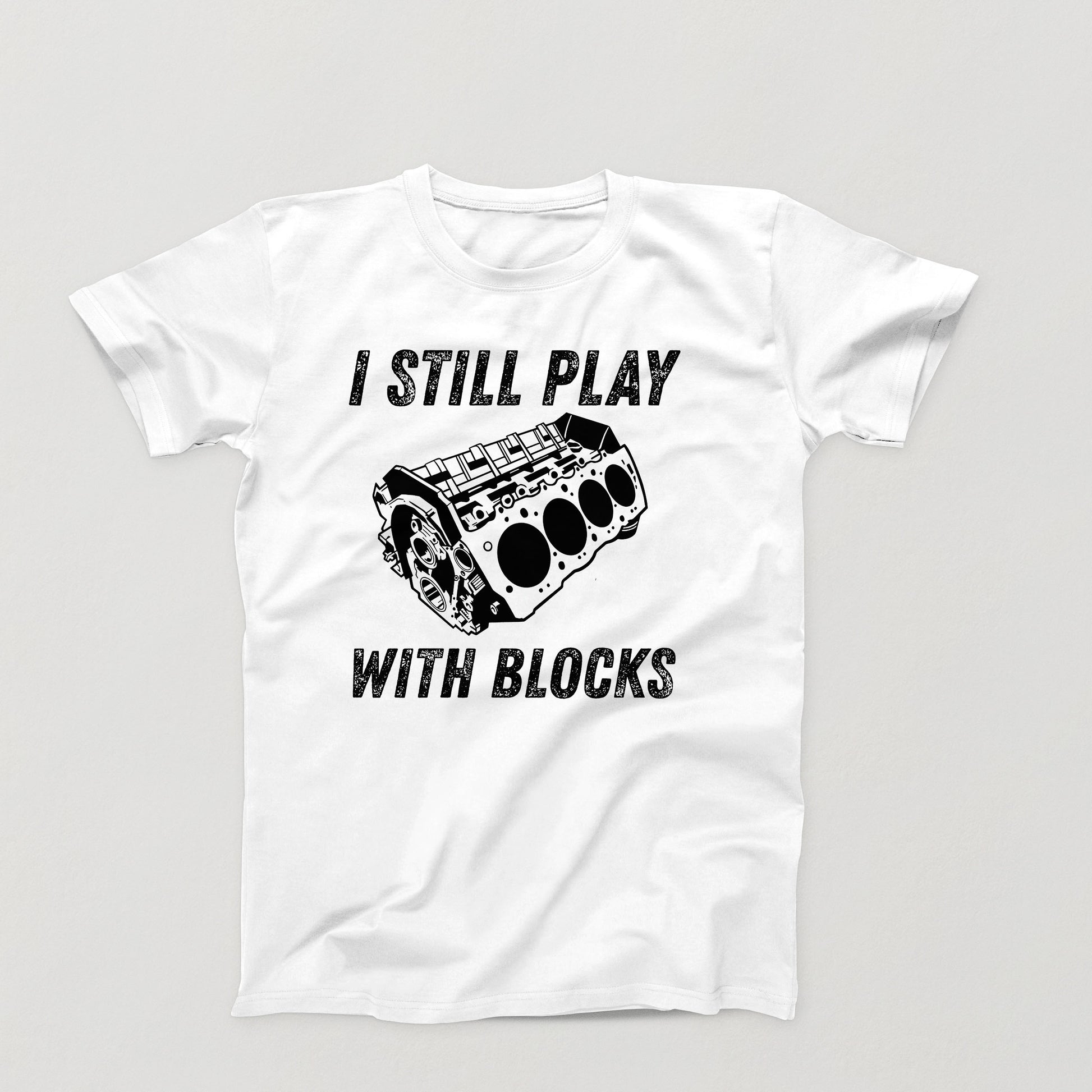 white t-shirt design with white V8 engine block and the phrase “I Still Play With Blocks” in distressed text