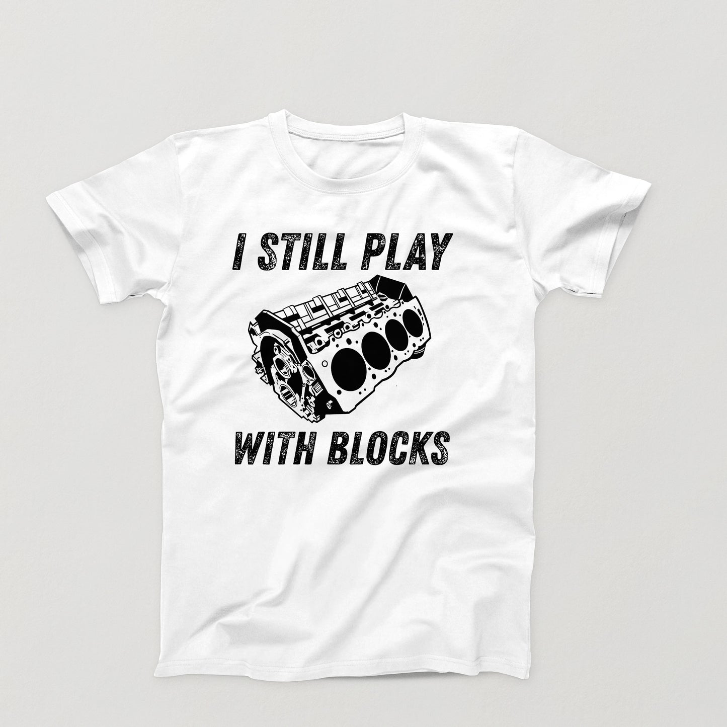 white t-shirt design with white V8 engine block and the phrase “I Still Play With Blocks” in distressed text